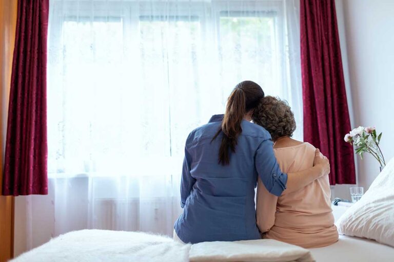 An adult child gently helping an older parent stand up from a chair in a cozy living room.